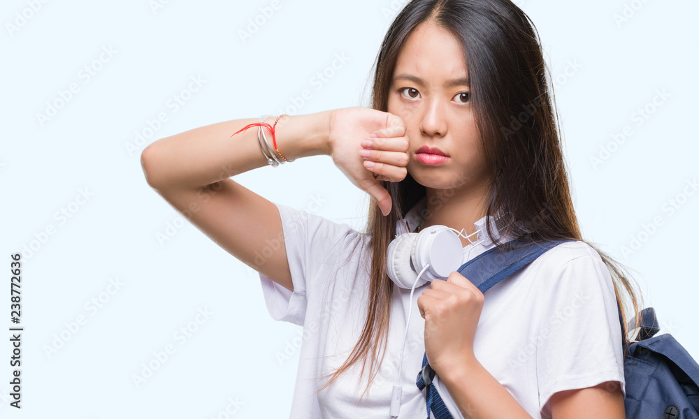 Young asian woman wearing backpack and headphones over isolated background with angry face, negative sign showing dislike with thumbs down, rejection concept Young asian woman wearing backpack and headphones over isolated background with angry face, negative sign showing dislike with thumbs down, rejection concept