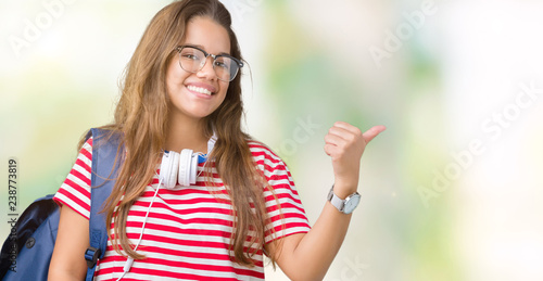 Young beautiful brunette student woman wearing headphones and backpack over isolated background smiling with happy face looking and pointing to the side with thumb up. © Krakenimages.com