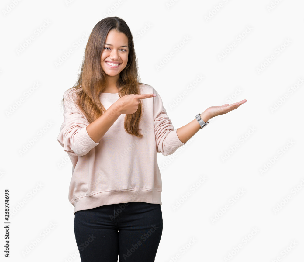 Young beautiful brunette woman wearing pink sweatshirt over isolated background amazed and smiling to the camera while presenting with hand and pointing with finger. Young beautiful brunette woman wearing pink sweatshirt over isolated background amazed and smiling to the camera while presenting with hand and pointing with finger.