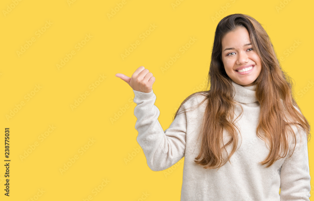 Young beautiful brunette woman wearing turtleneck sweater over isolated background smiling with happy face looking and pointing to the side with thumb up. Young beautiful brunette woman wearing turtleneck sweater over isolated background smiling with happy face looking and pointing to the side with thumb up.