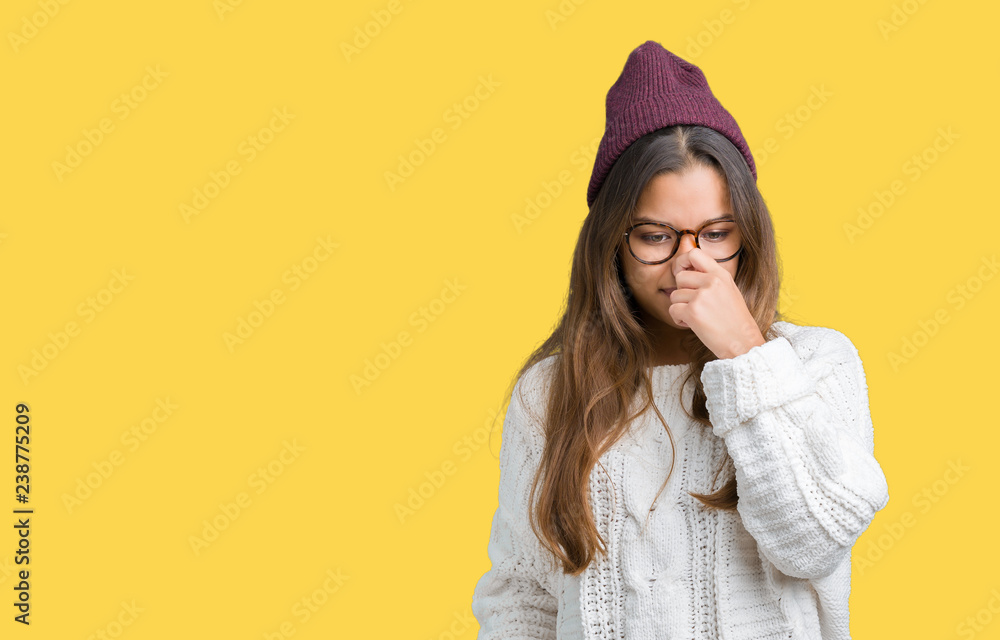 Young beautiful brunette hipster woman wearing glasses and winter hat over isolated background smelling something stinky and disgusting, intolerable smell, holding breath with fingers on nose Young beautiful brunette hipster woman wearing glasses and winter hat over isolated background smelling something stinky and disgusting, intolerable smell, holding breath with fingers on nose