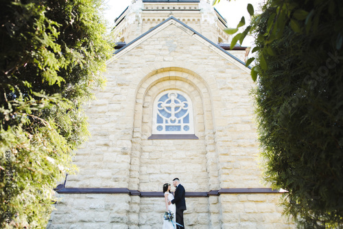 Stunning and happy wedding couple poses on the green lawn before an old church