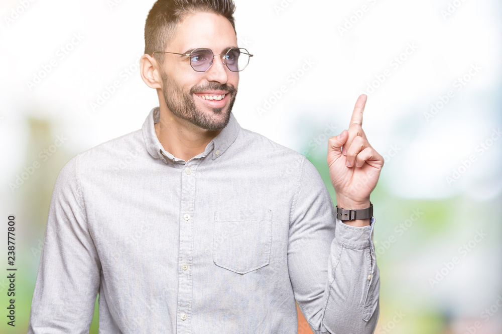 Young handsome man wearing sunglasses over isolated background with a big smile on face, pointing with hand and finger to the side looking at the camera. Young handsome man wearing sunglasses over isolated background with a big smile on face, pointing with hand and finger to the side looking at the camera.