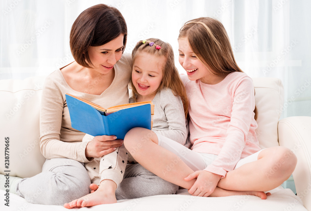 Mom and her two little daughters reading children's book in the living room. Mom and daughters sitting on the coach and reaading Mom and her two little daughters reading children's book in the living room. Mom and daughters sitting on the coach and reaading