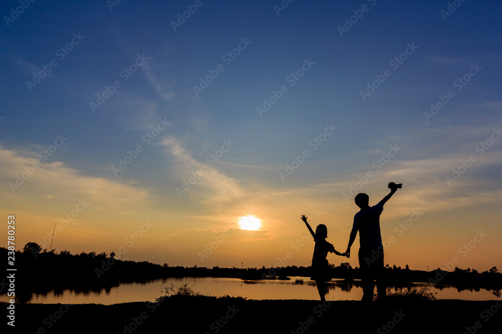 silhouette of father and son stood holding hands arms lake on sunset.