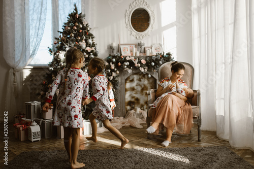 Two little sisters in pajamas having fun New Year's tree with gifts in the light cozy room and their mother sits in the armchair with little baby next to the fireplace