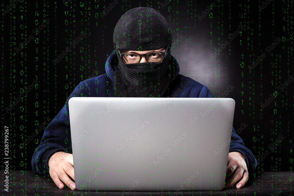binary code on the background of a cybercriminal in a balaclava and glasses sits at a laptop binary code on the background of a cybercriminal in a balaclava and glasses sits at a laptop