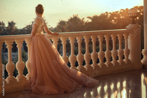 Beautiful lady in luxurious ballroom dress with tulle skirt and lacy top standing on the large balcony looking away at sunset. Back view. Text space