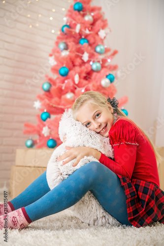 little blonde girl sits in a bright room on the floor against the background of a pink Christmas tree and hugs a pillow
