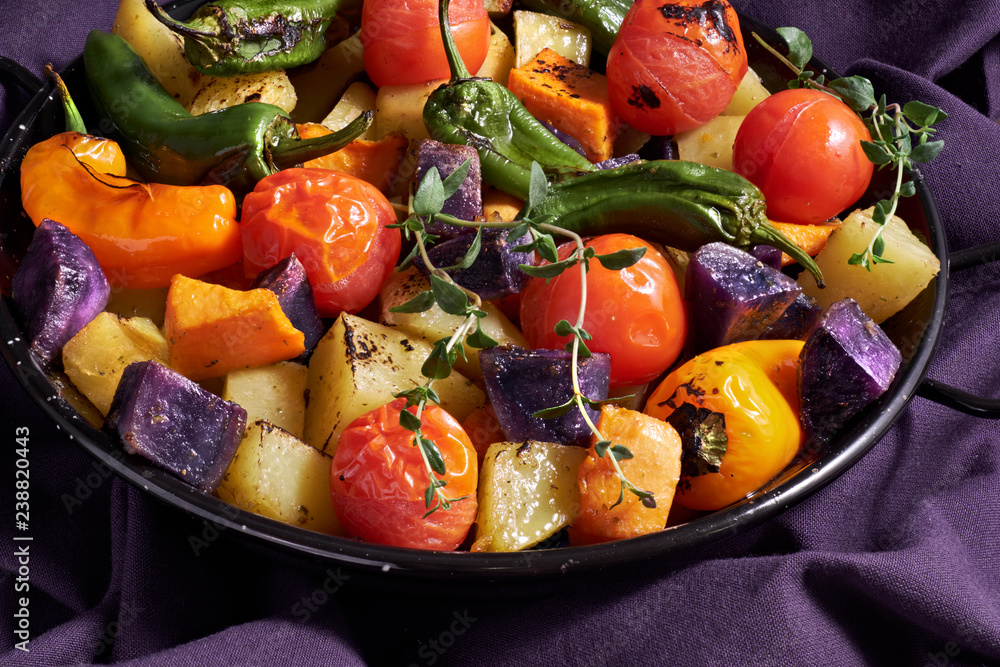 Rustic, oven baked vegetables in baking dish. Seasonal vegetarian vegan meal on dark purple linen towel