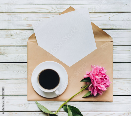 A white cup of coffee stands next to a faded peonys and an open kraft envelope containing a white sheet of paper. Copy space, top view.