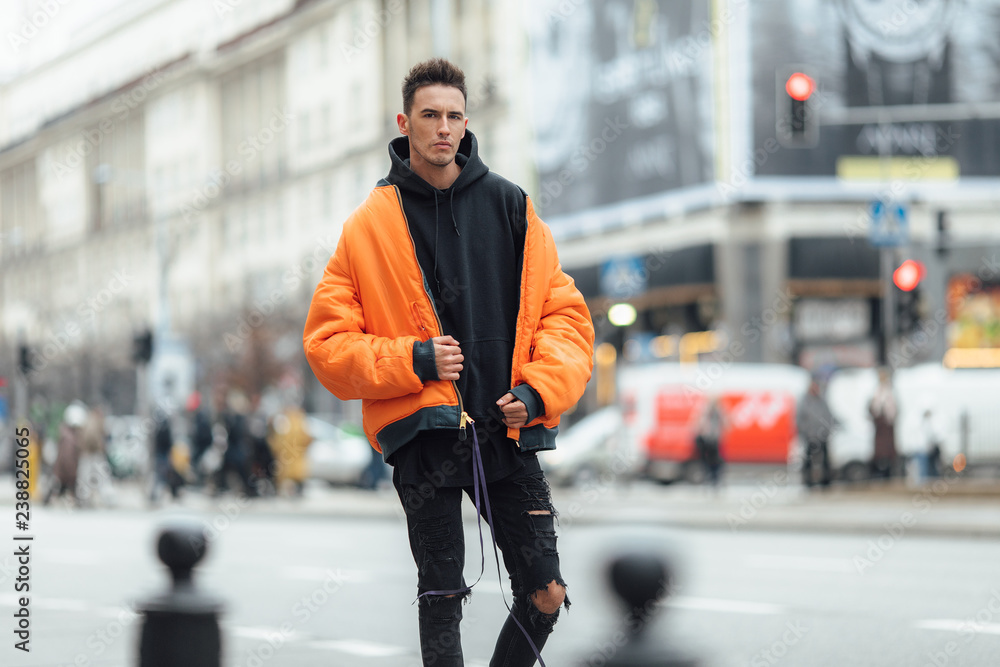 Fashionable man stand on the street near buildings. Wear orange jacket and all black. Winter, autumn outfit. Jacket with blouse and black sneakers.