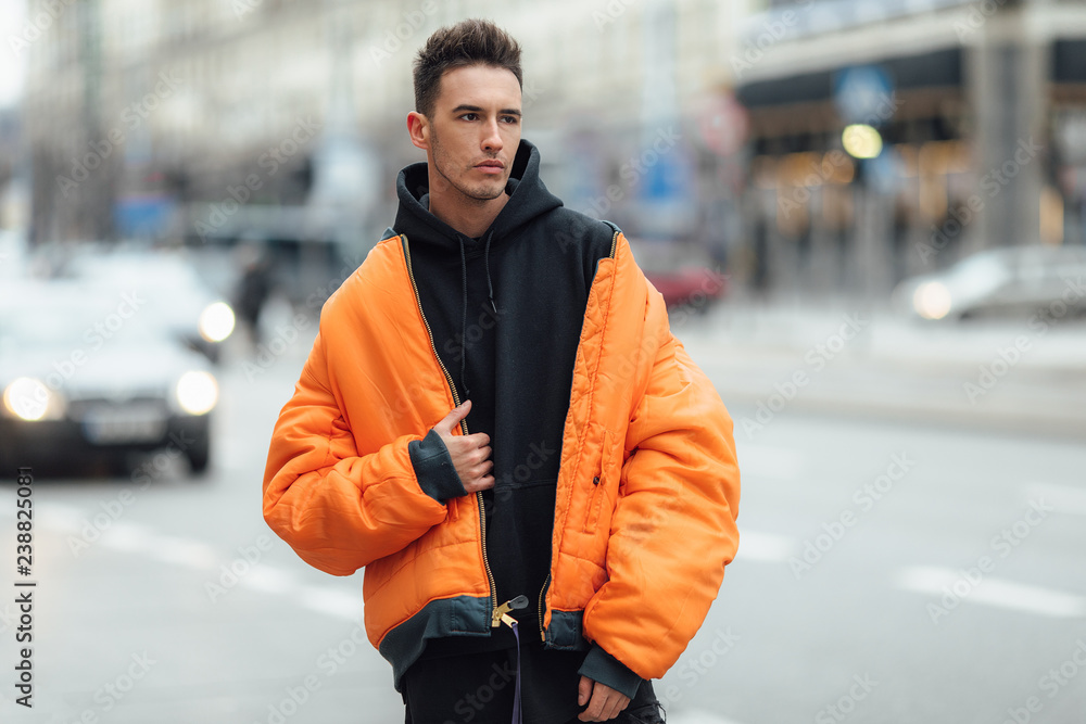 Fashionable man stand on the street near buildings. Wear orange jacket and  all black. Winter, autumn outfit. Jacket with blouse and black sneakers.  Photos | Adobe Stock