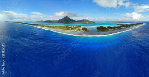 Aerial panoramic landscape view of the island of Bora Bora in French Polynesia with the Mont Otemanu mountain surrounded by a turquoise lagoon, motu atolls, reef barrier, and the South Pacific Ocean