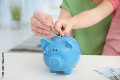 Little girl with grandmother putting money into piggy bank at table, closeup