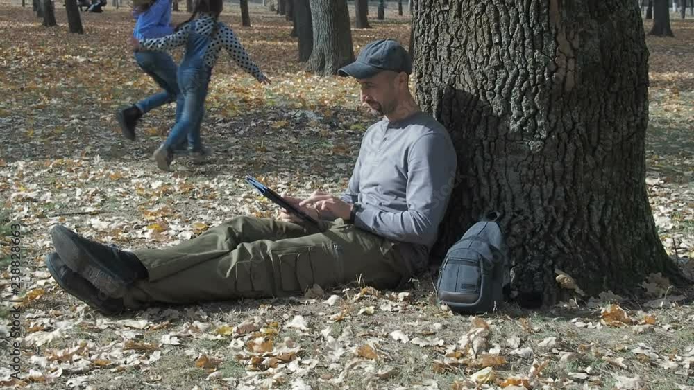 Family on vacation. A man sits with a tablet under a tree in an autumn park, and his fly is playing nearby. Family on vacation. A man sits with a tablet under a tree in an autumn park, and his fly is playing nearby.
