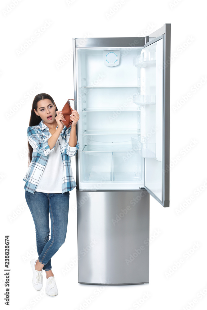 Young woman with no money in wallet near empty refrigerator on white background Young woman with no money in wallet near empty refrigerator on white background