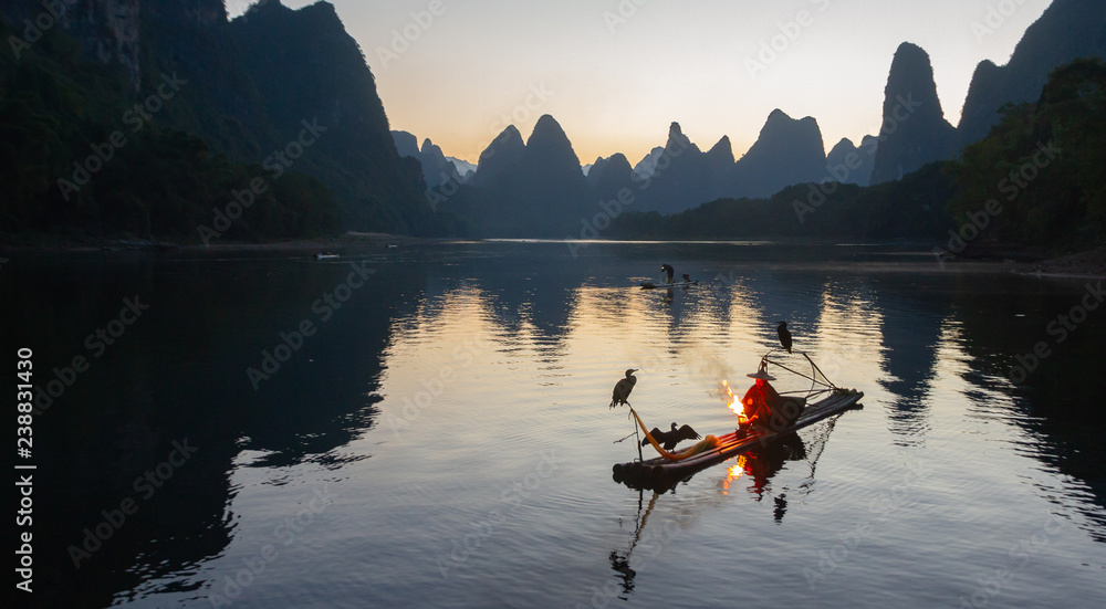 Cormorant fisherman on raft in lake in Guilin, China, with three cormorant birds. Fisherman is using a bright flame to heat teapot and light pipe. - obrazy, fototapety, plakaty 