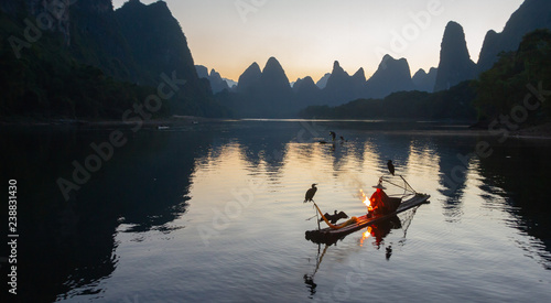 Cormorant fisherman on raft in lake in Guilin, China, with three cormorant birds. Fisherman is using a bright flame to heat teapot and light pipe.