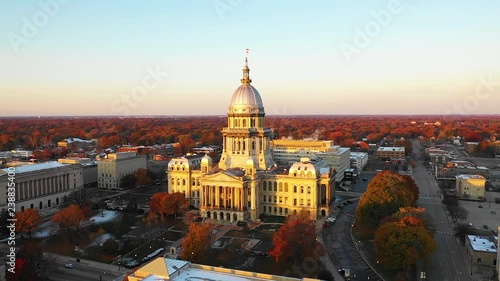 Stunning slow motion establishing aerial drone shot of the Illinois State Capitol Building in Springfield, Illinois, at dawn in November as the sun rises and fall leaves glow orange in the distance. photo