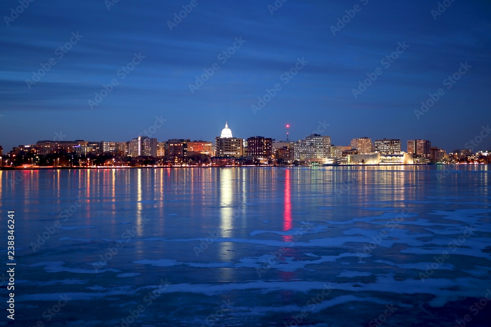 Madison downtown skyline illuminated at winter night with official buildings, Monona Terrace and capitol dome, glowing in the dark. Cityscape reflects in a frozen lake Monona.