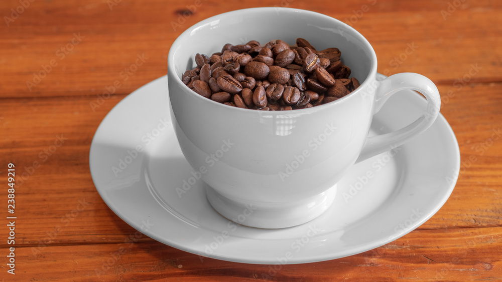 Cup of roasted coffee beans in white ceramic cup, on small white ceramic plate, on wooden surface Cup of roasted coffee beans in white ceramic cup, on small white ceramic plate, on wooden surface