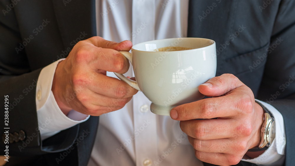 Man holding a cup of coffee with cream in white ceramic cup, dressed in white dress shirt and black business suit and wearing a watch Man holding a cup of coffee with cream in white ceramic cup, dressed in white dress shirt and black business suit and wearing a watch