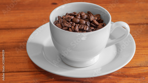 Cup of roasted coffee beans in white ceramic cup  on small white ceramic plate  on wooden surface