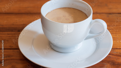 Cup of coffee with cream in white ceramic cup, on small white ceramic plate, on wooden surface