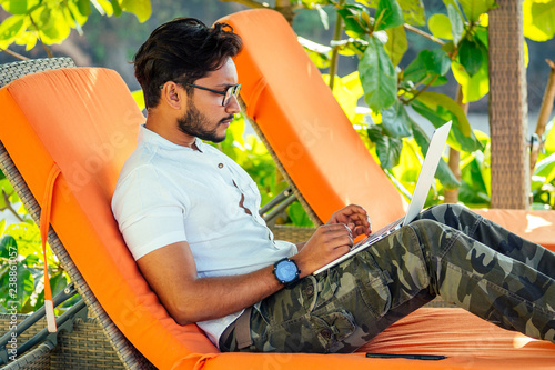 Canvas Print handsome and successful indian man in a stylish well-dressed freelancer working with a laptop on the beach