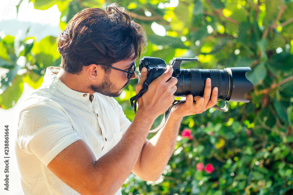 Fototapeta premium handsome and confident indian man photographer with a large professional camera taking pictures photo shooting on the beach.photo session on summer holiday on the background of green tropical trees