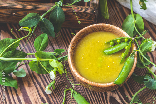 Wooden plate of pea soup and pods of fresh green peas on dark wooden surface.