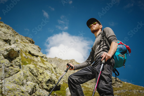 Porter of a stylish hipster traveler with a beard and a backpack in sunglasses and a cap with trekking poles standing on a rock against the background of a slope and blue sky in the mountains