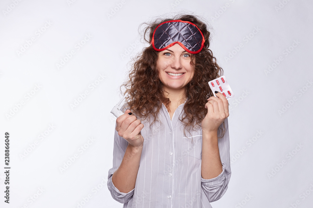 Fototapeta premium Pretty Caucasian female woke up early morning, wearing pajamas and sleeping mask, looks happy and stylish. Beautiful young woman realize that she is healthy and do not need medicine and pills anymore.