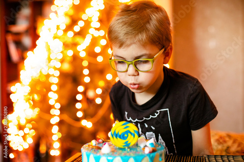 Beautiful adorable seven eight year old boy in grey shirt, celebrating his birthday, blowing candles on homemade baked cake, indoor photo