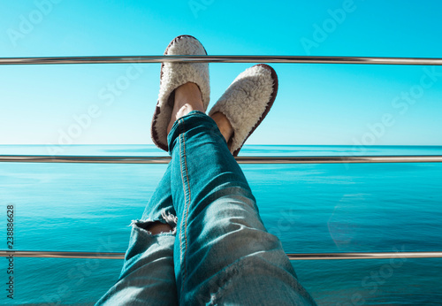 Women's legs in torn jeans and white sheep fur slippers lying on the crossbar of the balcony against the backdrop of the blue endless sea and sky early summer morning. Beautiful relaxing background.