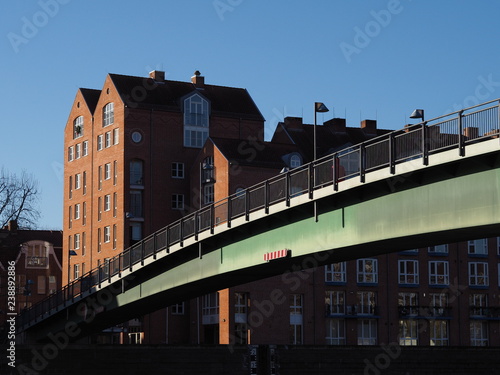 Bremen, Germany - Steel bridge across the river Weser leading to the Teerhof peninsula with red brick buildings in the background under a clear blue sky © Harald