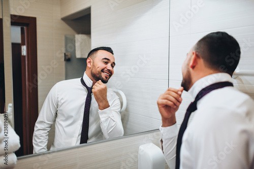 Young and stylish groom with a beard looks in the mirror. Morning groom. Wedding interior. Fees groom in the room. Wedding concept. Wedding photography.