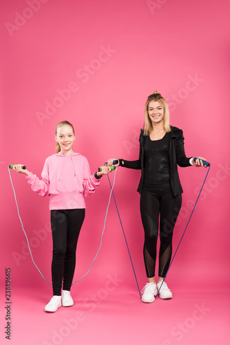 sportive mother and daughter in sportswear posing with skipping ropes, isolated on pink
