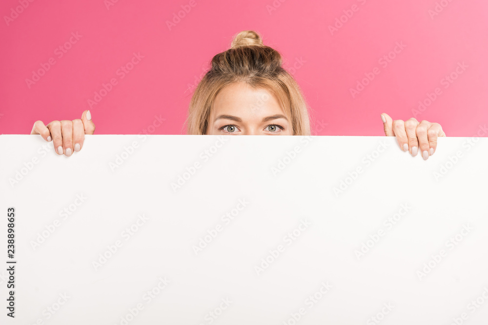 blonde woman hiding behind empty board, isolated on pink blonde woman hiding behind empty board, isolated on pink