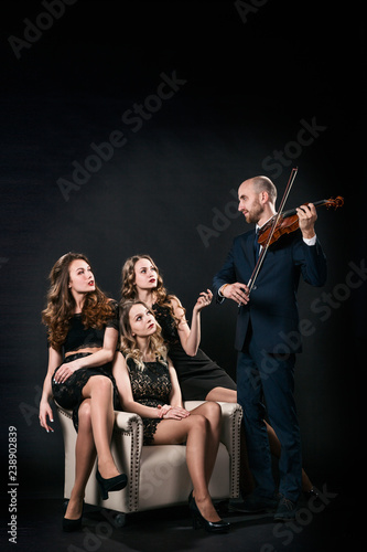 Three beautiful girls in black evening dresses are sitting in the chair, and the man plays the violin