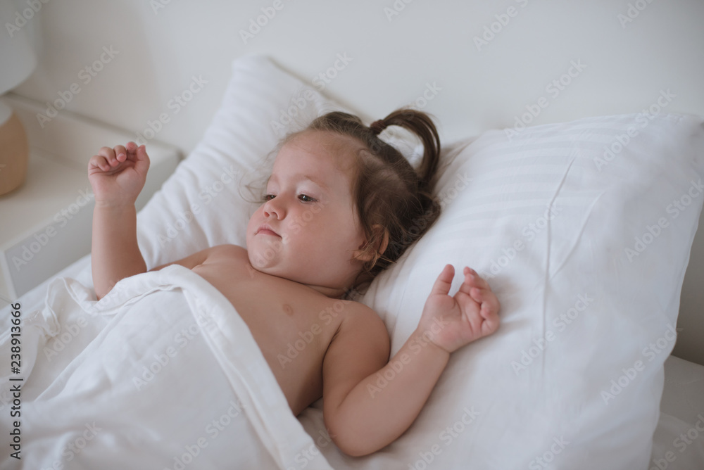 A little pretty dark-haired toddler girl is lying in bed smiling and looking happy. A little pretty dark-haired toddler girl is lying in bed smiling and looking happy.