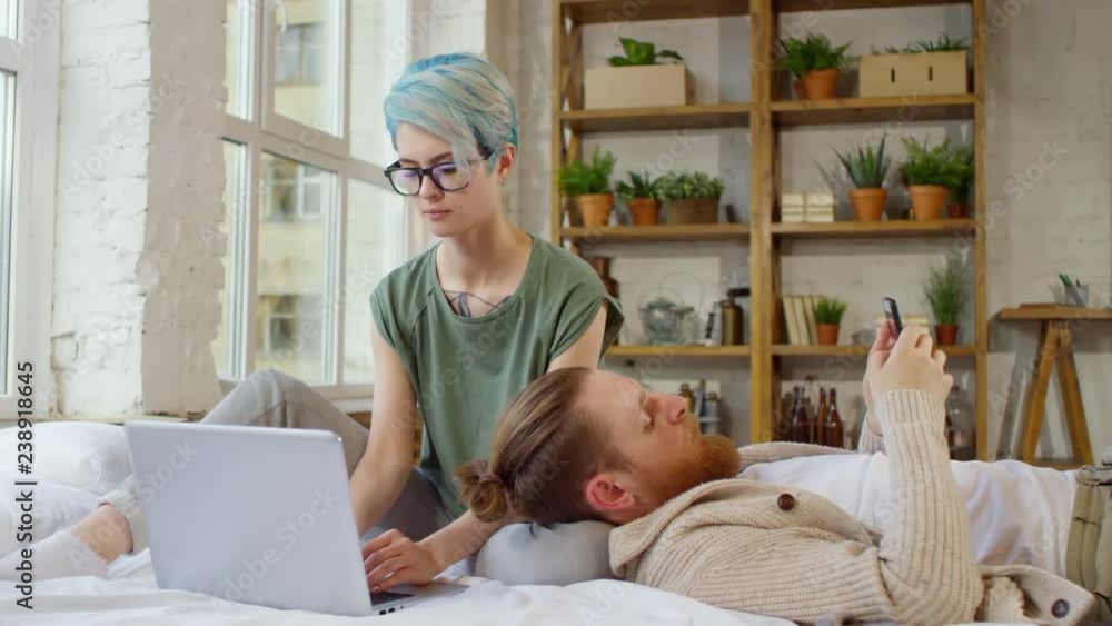 Tracking shot of young woman with blue hair and glasses sitting on bed and working on laptop as her bearded boyfriend lying beside her and browsing social media on mobile phone Tracking shot of young woman with blue hair and glasses sitting on bed and working on laptop as her bearded boyfriend lying beside her and browsing social media on mobile phone