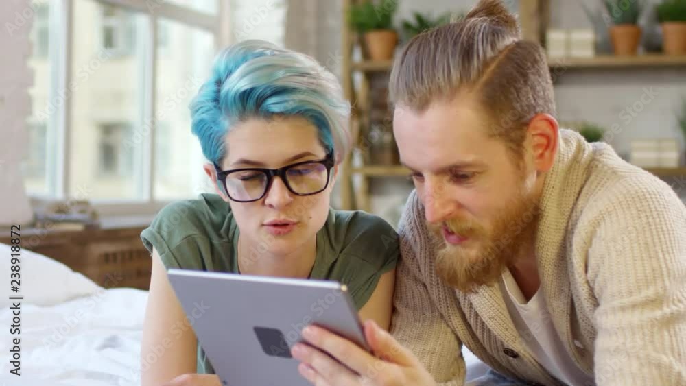 Tracking shot of young hipster woman with blue hair and man with beard lying on bed and chatting while watching something on tablet Tracking shot of young hipster woman with blue hair and man with beard lying on bed and chatting while watching something on tablet