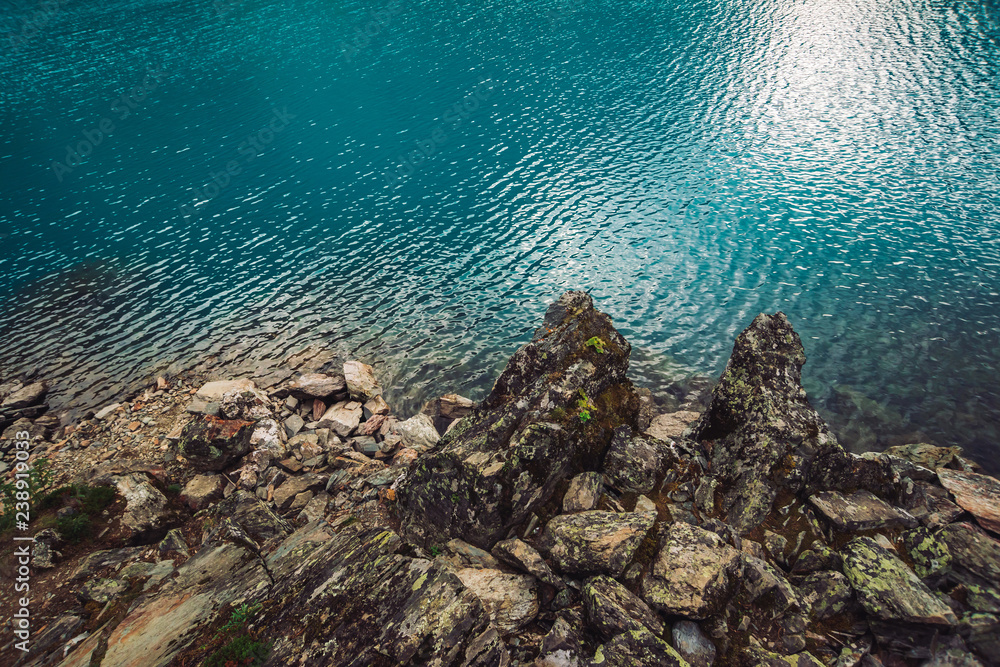 Rocky shore. Water edge. Shiny surface of azure mountain lake. Stony bottom in transparent water. Minimalist blue background. Reflection of mountains on clear water. Vegetation on stones. Copy space.
