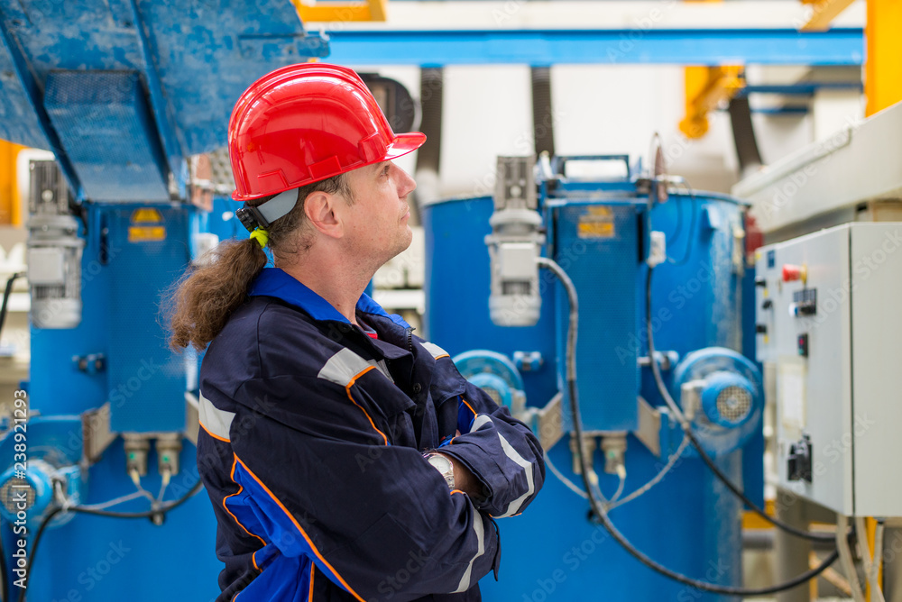 Worker in a factory wearing blue working suit and red helmet looking up in machines in the background Worker in a factory wearing blue working suit and red helmet looking up in machines in the background