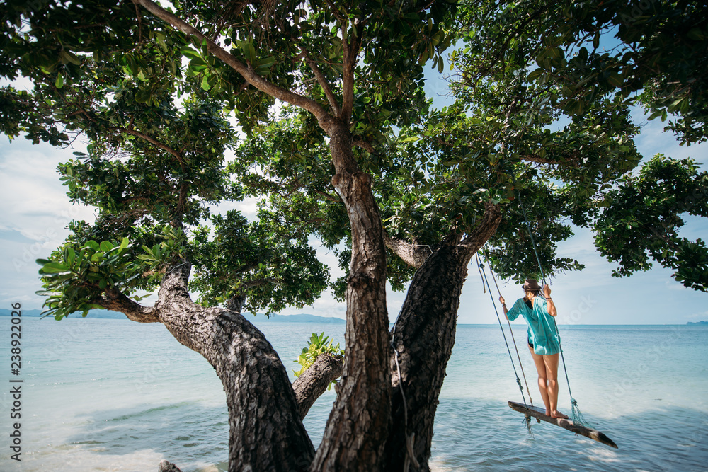 A young woman is swinging on a swing in the shade of a big tree by the sea. Girl on the beach swing on the coast of a tropical island.