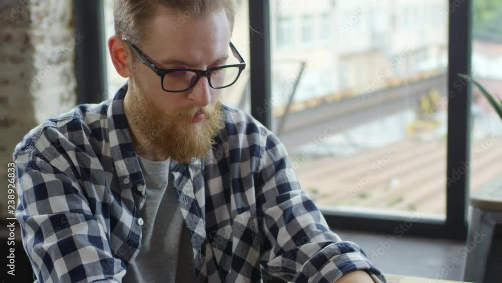 Tilt down shot of young man with beard and glasses sitting at desk by window and working on laptop Tilt down shot of young man with beard and glasses sitting at desk by window and working on laptop