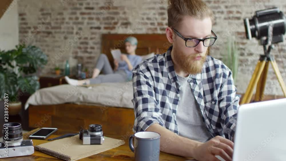 Medium shot of young hipster man with beard and glasses sitting at desk and working on laptop. Woman with blue hair relaxing on bed and reading book in background Medium shot of young hipster man with beard and glasses sitting at desk and working on laptop. Woman with blue hair relaxing on bed and reading book in background