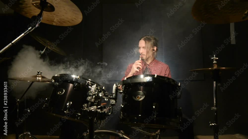 A long-haired drummer sitting behind a drum kit is hovering with a vape. Vape culture in music. Static plan A long-haired drummer sitting behind a drum kit is hovering with a vape. Vape culture in music. Static plan